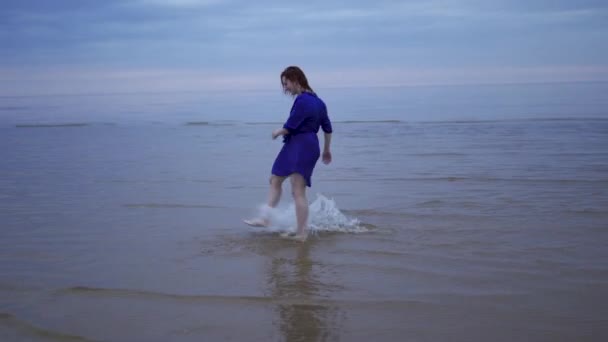 Молодая рыжая девушка в синем платье с мокрыми волосами, идущими по воде и делающими брызги - Настроенный холодный закат с яркими пурпурно-голубоватыми красками в заливе Балтийского моря летом — стоковое видео