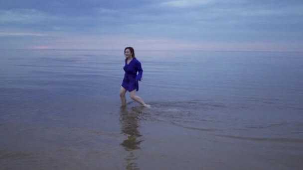 Νεαρή κοκκινομάλλα κοπέλα με μπλε φόρεμα με βρεγμένα μαλλιά να περπατά πάνω από το νερό και να κάνει πιτσιλιές - Moody κρύο ηλιοβασίλεμα με ζωηρά μωβ χρώματα στον Κόλπο της Βαλτικής το καλοκαίρι — Αρχείο Βίντεο