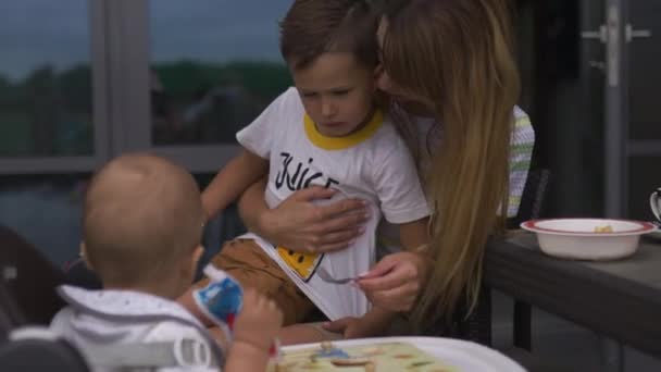 Молода мама годує свого сина хлопчика сидячи на дитячому сидінні - Сім'я цінує теплий колір літньої сцени — стокове відео