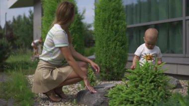 Genç anne oynarken ve yeşil bir bahçede onun bebek erkek erkek kardeşi ile eğlenmek - Aile değerleri sıcak renk yaz sahnesi - Yeşil çiçek çalılar