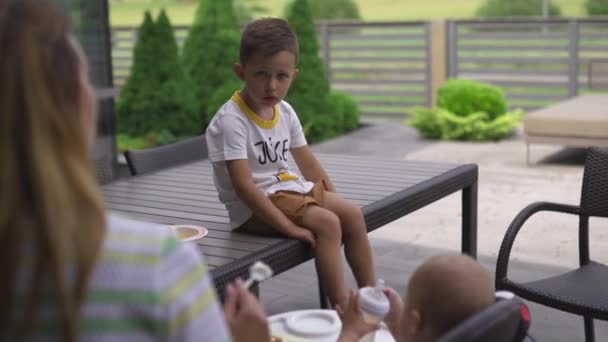 Irmão mais velho assistindo: Mãe jovem alimentando seu filho menino sentado em um assento de criança - Família valoriza cor quente cena de verão — Vídeo de Stock