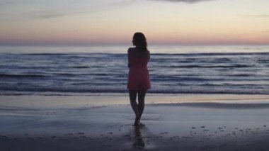 Açık kırmızı elbiseli genç kadın suyun üzerinde duruyor. Hayatın ve doğanın tadını çıkarıyor. Günbatımında uzak bir arka planda.