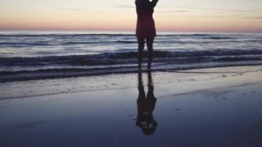 Açık kırmızı elbiseli genç kadın suyun üzerinde duruyor. Hayatın ve doğanın tadını çıkarıyor. Günbatımında uzak bir arka planda.