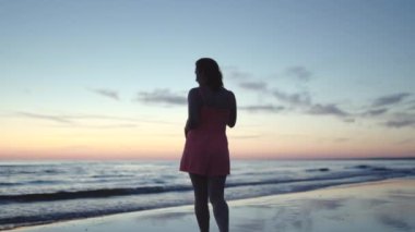 Açık kırmızı elbiseli genç kadın suyun üzerinde duruyor. Hayatın ve doğanın tadını çıkarıyor. Günbatımında uzak bir arka planda.