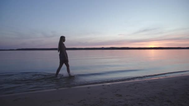 Młoda kobieta w krótkiej letniej sukni spacerowej wzdłuż plaży ciesząc się życiem i naturą daleko w tle na zachodzie słońca — Wideo stockowe