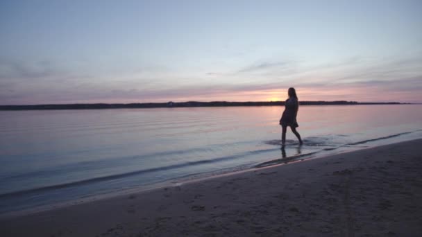 Junge Frau im kurzen Sommerkleid spaziert am Strand entlang und genießt das Leben und die Natur weit entfernt im Hintergrund bei Sonnenuntergang — Stockvideo