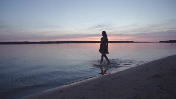 Młoda kobieta w krótkiej letniej sukni spacerowej wzdłuż plaży ciesząc się życiem i naturą daleko w tle na zachodzie słońca — Wideo stockowe