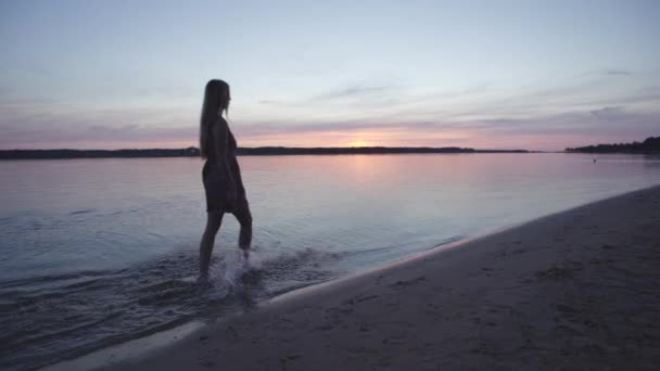 Mujer joven en un vestido corto de verano caminando y salpicando a lo largo de la playa disfrutando de la vida y la naturaleza fondo lejano en el atardecer — Vídeos de Stock