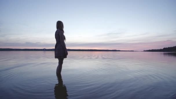 Jovem mulher em um vestido de verão curto em pé perto do rio desfrutando de vida e natureza fundo distante no pôr do sol — Vídeo de Stock