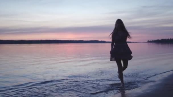 Jonge vrouw in een korte zomer jurk dansen langs op het water genieten van het leven en de natuur ver weg achtergrond bij de zonsondergang — Stockvideo