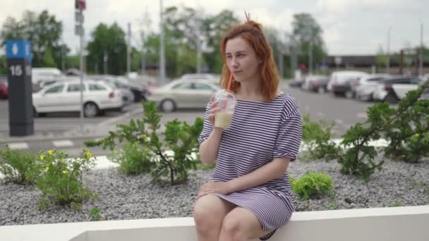 Jovem mulher ruiva feliz sentado e bebendo tirar café frio vestindo luz vestido listrado no verão com carros e folhas verdes no fundo — Vídeo de Stock