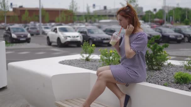 Счастливая молодая рыжая женщина сидит и пьет забрать холодный кофе носить легкие полосатые платья летом с автомобилями и зеленые листья на заднем плане — стоковое видео