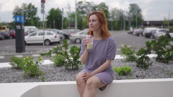 Jovem mulher ruiva feliz sentado e bebendo tirar café frio vestindo luz vestido listrado no verão com carros e folhas verdes no fundo — Vídeo de Stock