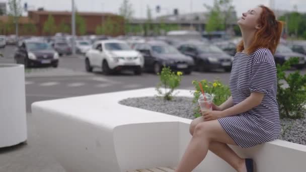 Jovem mulher ruiva feliz sentado e bebendo tirar café frio vestindo luz vestido listrado no verão com carros e folhas verdes no fundo — Vídeo de Stock