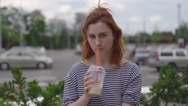 幸せな若い赤毛の女性が座って飲んで、車と緑の葉を背景に夏に軽いストライプのドレスを着て冷たいコーヒーを取り除きます — ストック動画