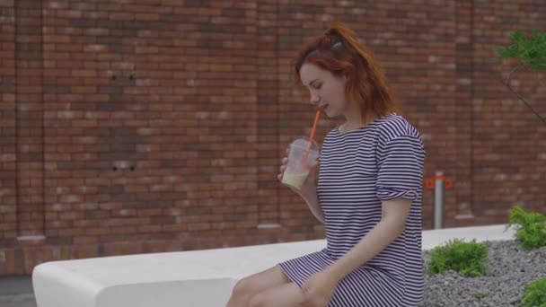 Счастливая молодая рыжая женщина пьет взять холодного кофе носить светлое полосатое платье летом с коричневой кирпичной стеной на заднем плане — стоковое видео