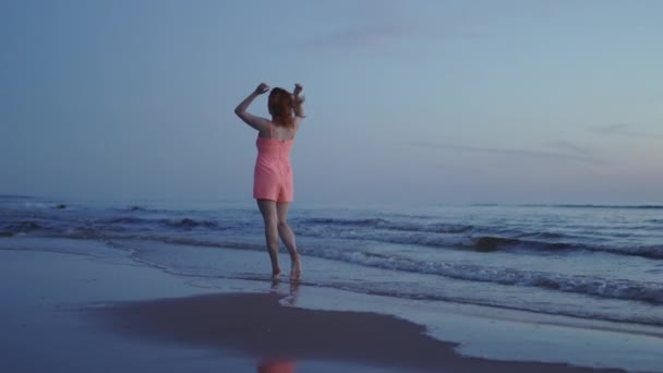 Junge Frau in leuchtend zartem roten Kleid, die am Strand entlang geht und das Leben und die Natur genießt, weit entfernt im Hintergrund bei Sonnenuntergang — Stockvideo