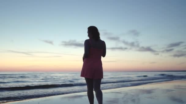 Νεαρή γυναίκα με φωτεινό τρυφερό κόκκινο φόρεμα στέκεται στο νερό απολαμβάνοντας τη ζωή και τη φύση πολύ μακριά φόντο στο ηλιοβασίλεμα — Αρχείο Βίντεο
