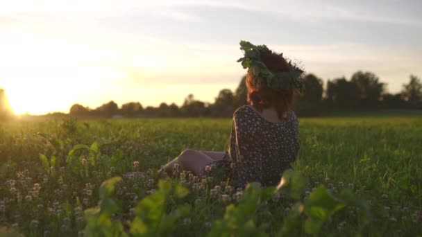 Zittend in gras: Redhead jonge vrouw in een krans tijdens de traditionele Letse Ligo Midzomerdag-blanke wit meisje dragen een gestippelde zomer jurk met een riem in zonnige zonsondergang — Stockvideo