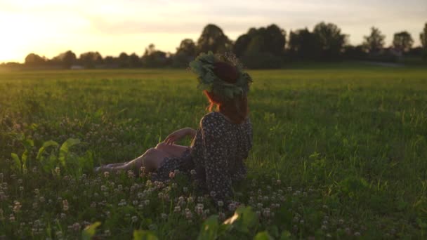 Çimde oturmak: Geleneksel Letonya Ligo yaz ortası sırasında çelenk redhead genç kadın - Beyaz beyaz kız güneşli günbatımında bir kemer ile noktalı bir yaz elbise giyiyor — Stok video
