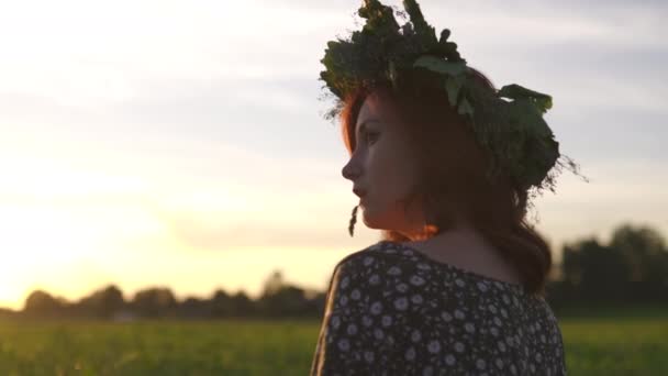 Assis dans l'herbe gros plan : jeune femme rousse dans une couronne lors de la traditionnelle journée de milieu de l'été Ligo letton - fille blanche caucasienne portant une robe d'été pointillée avec une ceinture au coucher du soleil ensoleillé — Video
