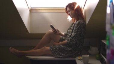 Mesajlaşma: Olumlu gülümseyen kızıl saçlı kadın teknoloji telefon kullanarak - Beyaz beyaz kız bir tavan arasında noktalı bir yazlık elbise giyiyor