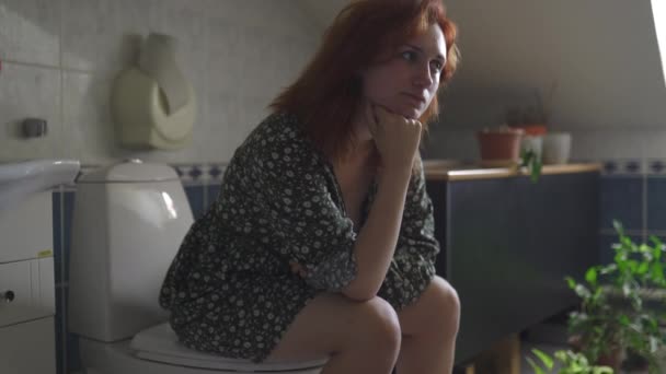 Mulher ruiva séria pensando em Significado da Vida sentado em um banheiro WC - menina branca caucasiana vestindo um vestido de verão pontilhado em um banheiro — Vídeo de Stock