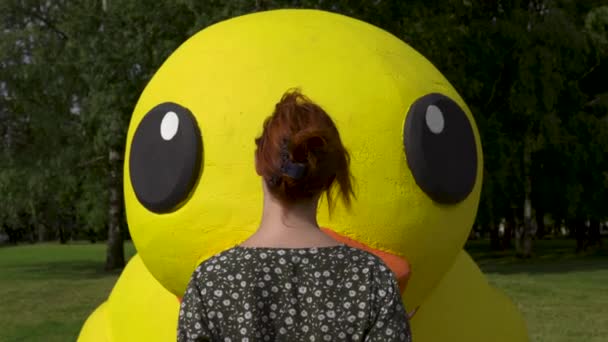 De pie: Mujer pelirroja y un enorme pato amarillo artificial no real o vivo - niña blanca caucásica con un vestido de verano punteado — Vídeos de Stock