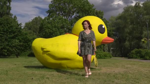 Serieus wandelen naar camera: redhead vrouw en een enorme kunstmatige niet echt of levend gele eend-Kaukasische wit meisje dragen een gestippelde zomer jurk — Stockvideo