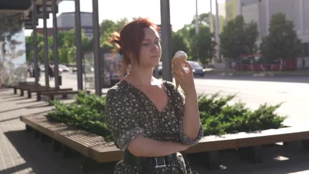 Turista pelirroja comiendo helado con coches y personas que pasan por - chica blanca caucásica con un vestido de verano punteado — Vídeo de stock