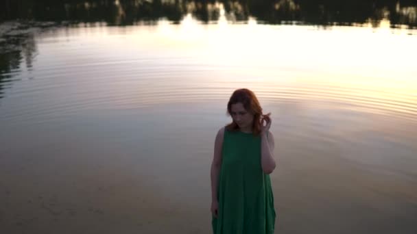 Mujer pelirroja de pie con un lago en el fondo reflejando el cielo puesta de sol — Vídeo de stock