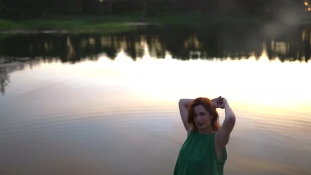 Mulher ruiva de pé com um lago no fundo refletindo céu pôr do sol — Vídeo de Stock