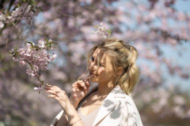 Bahar Sakura Cherry Blossom parkta güzel sarışın genç kadın onu seyahat turist serbest zaman sırasında doğa ve serbest zaman zevk - beyaz pantolon ve bej ceket ile t-shirt giyiyor