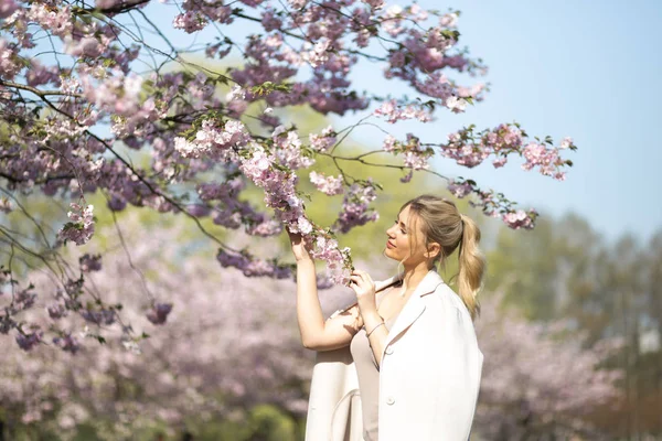 Jovem loira bonita no parque Sakura Cherry Blossom na primavera desfrutando da natureza e tempo livre durante o seu tempo livre de turista viajante - vestindo calças brancas e camiseta com uma jaqueta bege — Fotografia de Stock