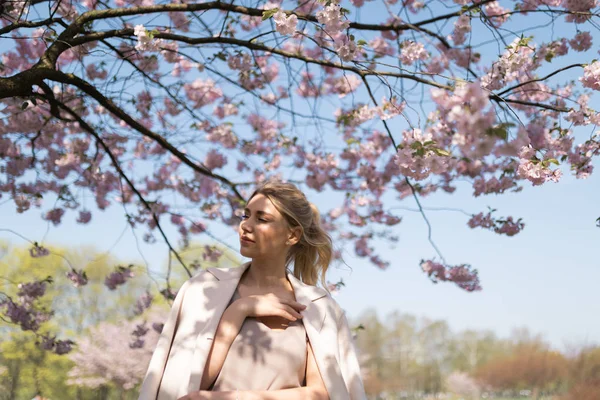 Jovem loira bonita no parque Sakura Cherry Blossom na primavera desfrutando da natureza e tempo livre durante o seu tempo livre de turista viajante - vestindo calças brancas e camiseta com uma jaqueta bege — Fotografia de Stock