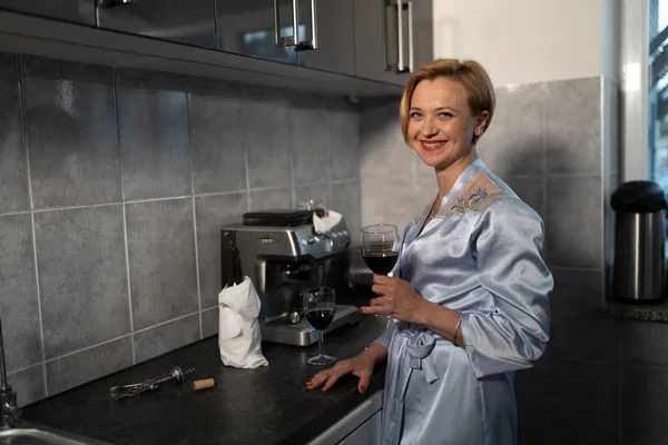 Svobodná žena pije červené víno ze sklenice v kuchyni v modrém ranním plášti-směje se a usmívá se — Stock fotografie