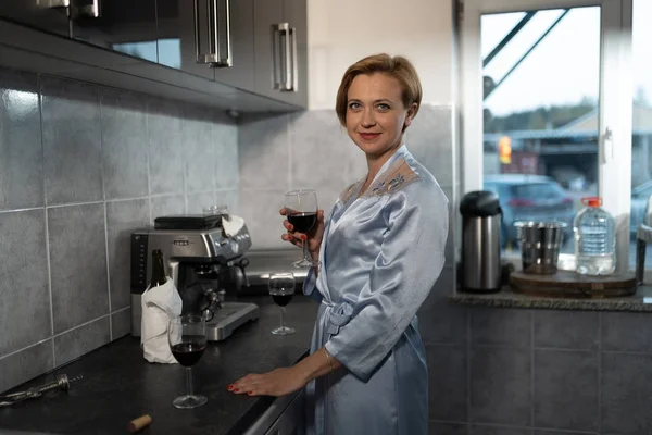 Single woman drinking red wine from a glass in a kitchen wearing blue morning gown - Laughing and smiling