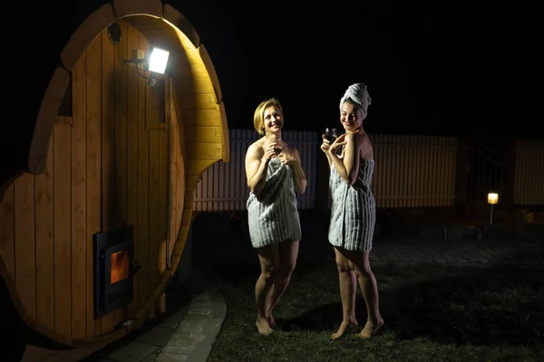 Two girlfriends women drinking red wine from glasses outsite near a wooden mobile sauna wearing just towels — Stock Photo, Image