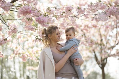 Genç anne düşen pembe yaprakları ve güzel çiçekleri ile çiçek açan Sakura Kiraz ağaçları altında onu küçük bebek erkek çocuk çocuk tutan