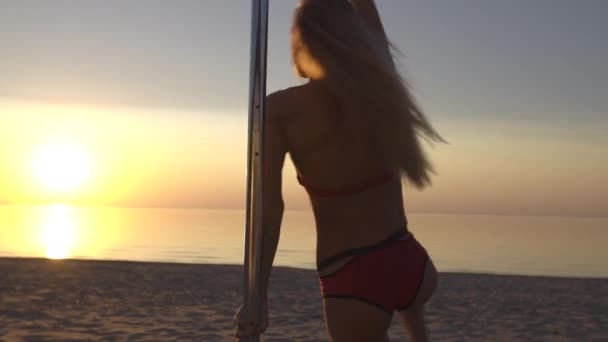 Κοντινό επάνω ευέλικτη θηλυκό πόλο χορεύτρια σε κόκκινα αθλητικά εσώρουχα φορούν άσκηση στον πυλώνα στο ηλιοβασίλεμα σε μια παραλία-δράκος τατουάζ και ακροβατικά γυμναστική — Αρχείο Βίντεο