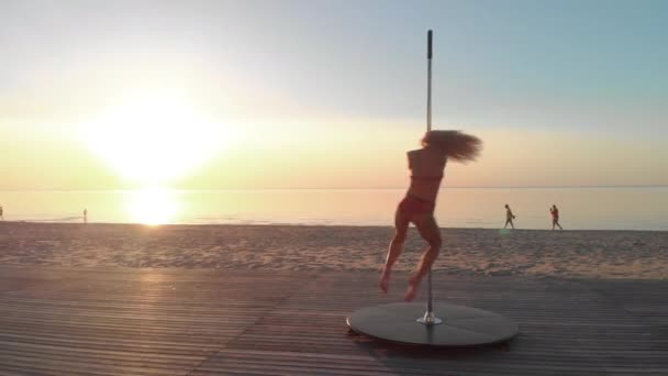 Aérea: Bailarina de polo flexible en lencería deportiva roja que hace ejercicio en el pilón al atardecer en una playa - Tatuaje de dragón y gimnasia acrobática — Vídeo de stock