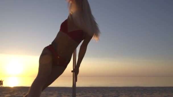 Close up Dançarina de pólo feminina flexível em roupas de lingerie esportiva vermelha exercitando-se no pilão ao pôr do sol em uma praia - Tatuagem de dragão e ginástica acromática — Vídeo de Stock