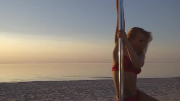 Fleksibel kvinnelig stripper i rødt sportsundertøy bruker treningsøvelse på pylonen ved solnedgang på en strand - Dragetatovering og akrobatisk gymnastikk – stockvideo