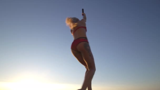 Bailarina femenina flexible en lencería deportiva roja que hace ejercicio en el pilón al atardecer en una playa - Tatuaje de dragón y gimnasia acrobática — Vídeos de Stock