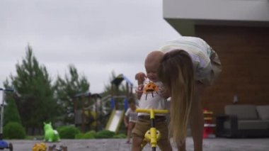 Genç anne oynarken ve bisiklet ile yeşil bir bahçede onun bebek erkek erkek kardeşi ile eğlenmek - Aile değerleri sıcak renk yaz sahnesi