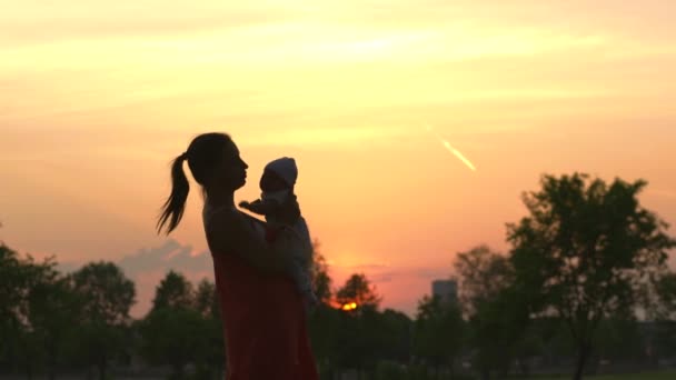 Sunset Silhouette: Joven madre sosteniendo a su bebé niño en el parque de la ciudad de pie frente al sol poniente y el cielo naranja vivo valores de la familia de color cálido escena de verano de mano — Vídeos de Stock