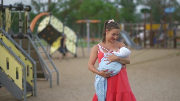 Breast feeding: Young mother breastfeeds her baby boy child in city park standing wearing bright red dress - Son wears white cap - Family values warm color summer scene handheld — Stock Video