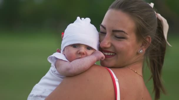 Mladá matka drží a hraje si s dětským dítětem v městském parku a má na sobě zářivě rudé šaty-syn nosí bílou čepici-rodinné hodnoty teplá barevná letní scéna handheld — Stock video