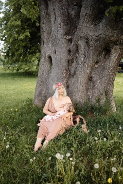 Emzirme: Genç hippi anne çiçek çerçeve daire giyen yeşil bir çim çim üzerinde bir ağacın altında oturan şehir parkında onun bebek kız çocuk emzirir