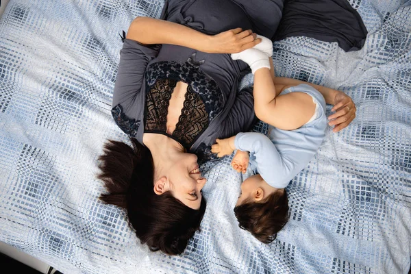 Madre embarazada acostada con su hijo bebé en la cama del dormitorio jugando y divirtiéndose - Niño asiático de etnia mixta con camisa de cuerpo azul con dibujos animados oso sonriente — Foto de Stock